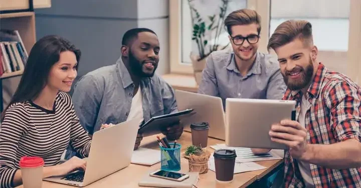 Casually dressed coworkers sit at a table with laptops and tablets as one member shows them something on his screen