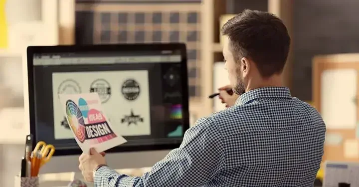 Graphic designer looking at paper that says "design" on it and logos on his computer screen